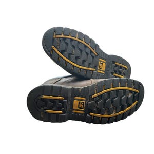 May include: Pair of brown work boots with black and yellow soles. The soles have a deep tread pattern for grip and feature the CAT logo. The boots are designed for durability and protection in demanding work environments.