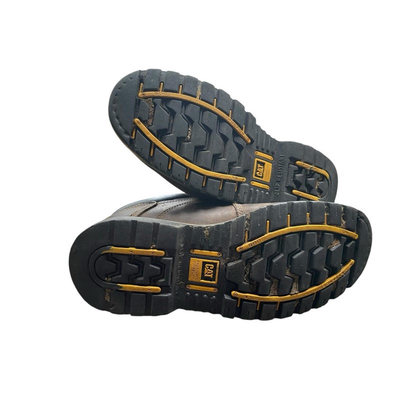 May include: Pair of brown work boots with black and yellow soles. The soles have a deep tread pattern for grip and feature the CAT logo. The boots are designed for durability and protection in demanding work environments.