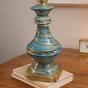 May include: A turquoise and gold ceramic table lamp, 10 inches tall. The lamp has a swirling design with gold accents and a brass lamp holder. It sits on a square base and is placed on a stack of books.
