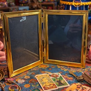 May include: A gold-colored, hinged picture frame with two empty black spaces. The frame is on a colorful patterned surface with tarot cards and jewelry. A lit candle and other decorative items are in the background.