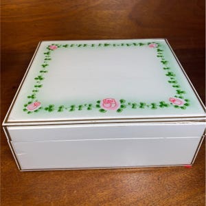 May include: A white jewelry box with a glass top featuring a floral design. The top has a border of green leaves and pink roses. The box has gold trim and a rectangular shape.