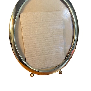 May include: Oval-shaped, gold-toned metal picture frame with a glass front. The frame has a simple design with a smooth, polished finish. The back of the frame is cardboard. The frame is supported by small, rounded feet.