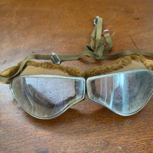 May include: Vintage aviator goggles with clear lenses and a brown frame. The goggles have a soft, furry lining and an olive green strap for securing them. The lenses are set in a metal frame.