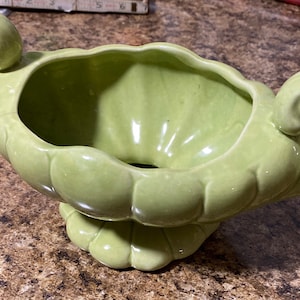 May include: A light green ceramic planter with two handles and a scalloped edge. The planter has a pedestal base.