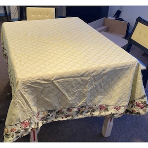 May include: A cream-colored tablecloth with a diamond pattern and a floral border. The tablecloth is made of a soft, woven fabric and has a subtle sheen.