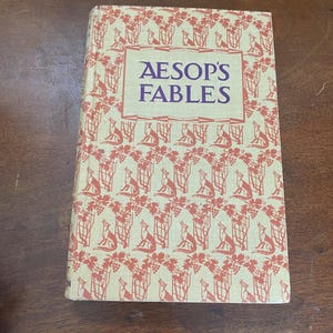 May include: A vintage hardcover book titled "Aesop's Fables." The book has a light beige cover with a repeating pattern of red foxes and grape vines. The title is in a purple rectangular box.