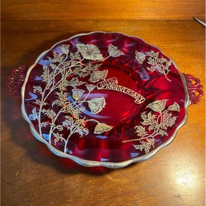 May include: A round ruby red glass serving platter with two side handles and a scalloped edge. The surface features a silver-colored floral pattern and a central banner with the text 40th ANNIVERSARY. The rim is trimmed in silver.