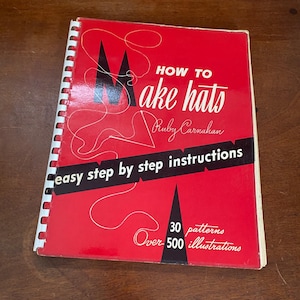 May include: A red book titled "How to Make Hats" by Ruby Carnahan. The cover features black geometric shapes and white text, including "easy step by step instructions." It contains 30 patterns and over 500 illustrations. The book is spiral-bound.