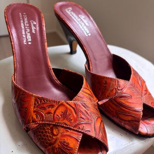 May include: A pair of orange peep-toe mules with a dark red interior lining. The exterior features an embossed floral pattern. The insole is printed with the text Couture Donald J Pliner Made in the mountains of Italy. The shoes have a mid-height heel.