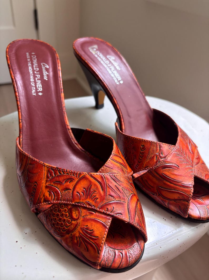 May include: A pair of orange peep-toe mules with a dark red interior lining. The exterior features an embossed floral pattern. The insole is printed with the text Couture Donald J Pliner Made in the mountains of Italy. The shoes have a mid-height heel.
