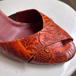 May include: An orange and reddish-brown peep-toe shoe featuring an embossed floral pattern across the upper surface. The shoe has a dark brown interior lining and a visible elastic strap detail near the opening, showcasing intricate texture.