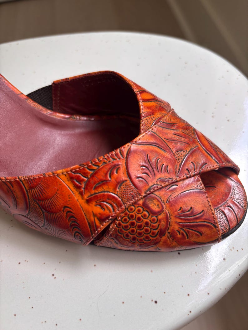 May include: An orange and reddish-brown peep-toe shoe featuring an embossed floral pattern across the upper surface. The shoe has a dark brown interior lining and a visible elastic strap detail near the opening, showcasing intricate texture.
