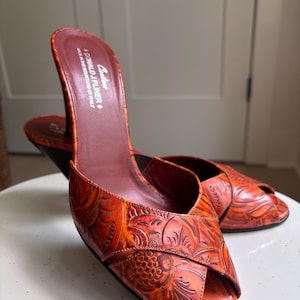 May include: A pair of orange peep-toe mules with a dark brown heel and an embossed floral pattern. The insole features the text "Couture DONALD J PLINER MADE IN THE MOUNTAINS OF ITALY". The shoes have a crossover strap design at the front.