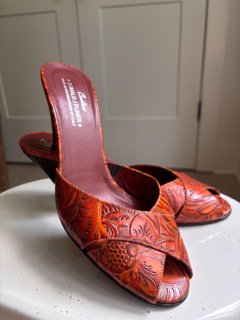 May include: A pair of orange peep-toe mules with a dark brown heel and an embossed floral pattern. The insole features the text "Couture DONALD J PLINER MADE IN THE MOUNTAINS OF ITALY". The shoes have a crossover strap design at the front.