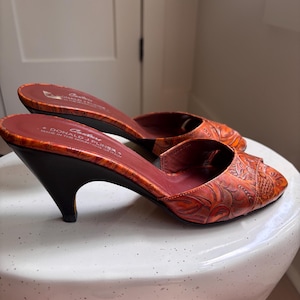 May include: A pair of orange peep-toe mules with a dark brown kitten heel and an embossed floral pattern. The interior footbed is dark red and features the text "Couture Donald J Pliner Made in the Mountains of Italy" printed in white.