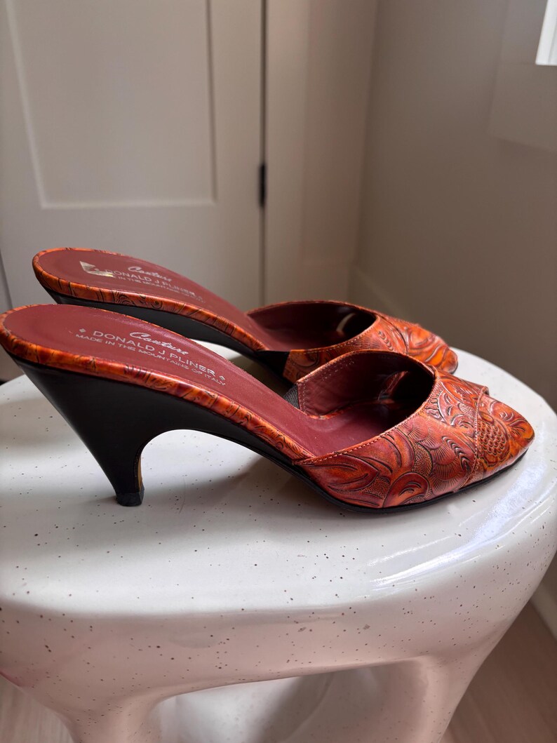 May include: A pair of orange peep-toe mules with a dark brown kitten heel and an embossed floral pattern. The interior footbed is dark red and features the text "Couture Donald J Pliner Made in the Mountains of Italy" printed in white.