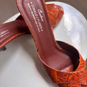 May include: A pair of orange open-toe mules with a textured floral pattern. The interior footbed is dark brown and features the text "Couture DONALD J PLINER MADE IN THE MOUNTAINS OF ITALY". The shoes have a slip-on design with a low heel.