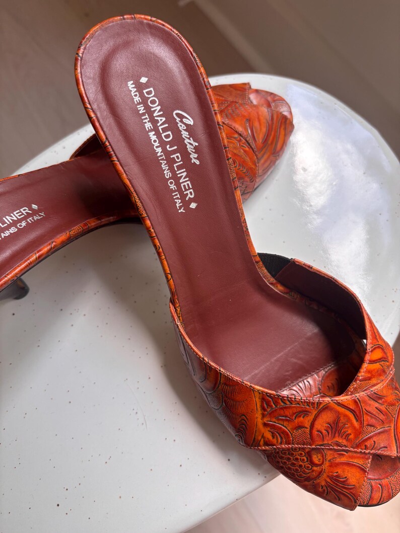 May include: A pair of orange open-toe mules with a textured floral pattern. The interior footbed is dark brown and features the text "Couture DONALD J PLINER MADE IN THE MOUNTAINS OF ITALY". The shoes have a slip-on design with a low heel.