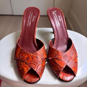 May include: A pair of orange and red embossed leather mules with an open-toe design and a mid-height heel. The interior footbed is dark maroon and features the text Donald J Pliner Couture Made in the Mountains of Italy printed in white.