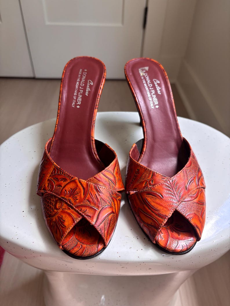 May include: A pair of orange and red embossed leather mules with an open-toe design and a mid-height heel. The interior footbed is dark maroon and features the text Donald J Pliner Couture Made in the Mountains of Italy printed in white.