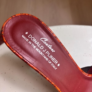 May include: The interior footbed of a shoe featuring a deep burgundy color. The text reads Couture DONALD J PLINER MADE IN THE MOUNTAINS OF ITALY. The edge of the footbed is trimmed with an orange and black patterned material with a textured finish.