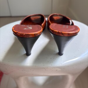 May include: A pair of open-toe mules featuring a textured orange and brown marbled pattern on the upper and footbed. The shoes have a dark brown kitten heel and a smooth brown interior lining with a visible brand logo printed on the heel area.