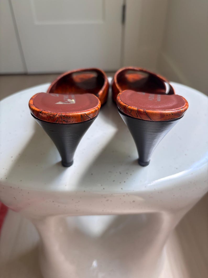 May include: A pair of open-toe mules featuring a textured orange and brown marbled pattern on the upper and footbed. The shoes have a dark brown kitten heel and a smooth brown interior lining with a visible brand logo printed on the heel area.