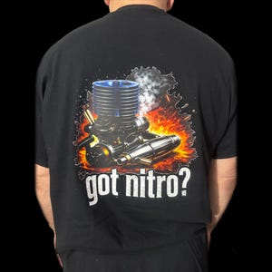 Puede incluir: Camiseta negra con un gráfico detallado de un motor de modelo con componentes azules y plateados, sobre un fondo de explosión de fuego. El texto "got nitro?" está impreso en blanco debajo.