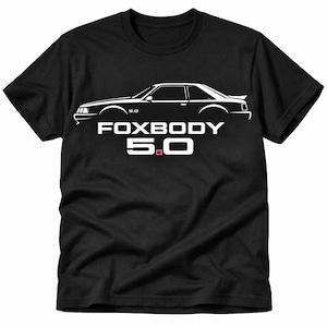 May include: Black t-shirt featuring a white graphic of a classic car's side profile. The text "FOXBODY 5.0" is displayed in white, with a red square in the "5.0". The shirt is made of a soft, comfortable material.