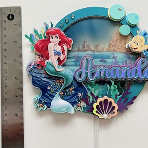 May include: A cake topper featuring Ariel, the Little Mermaid, sitting on a rock with a seashell and seaweed. The topper is decorated with a blue and green background and the name "Amando" in purple and blue lettering.