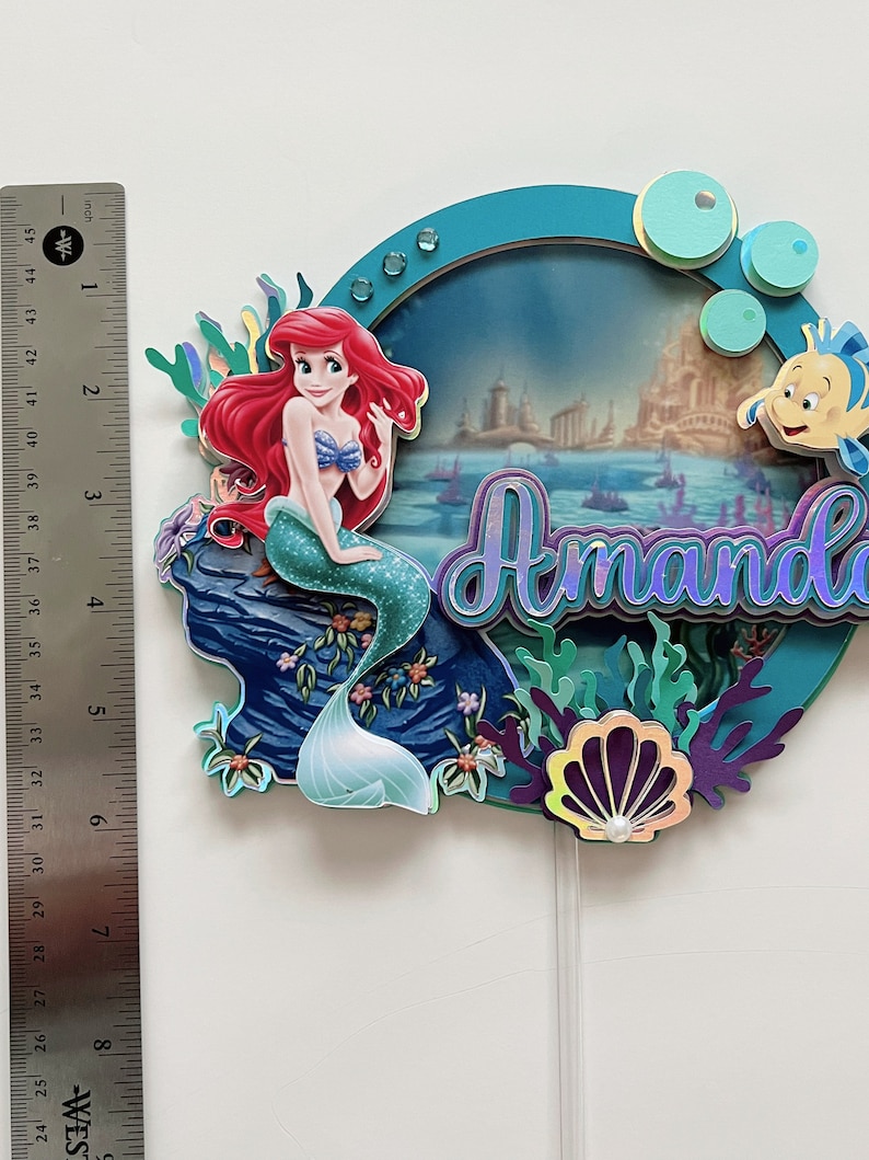 May include: A cake topper featuring Ariel, the Little Mermaid, sitting on a rock with a seashell and seaweed. The topper is decorated with a blue and green background and the name "Amando" in purple and blue lettering.