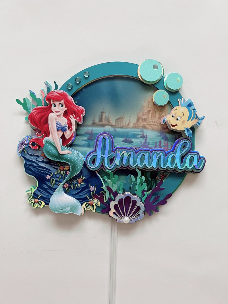 May include: A cake topper featuring Ariel, the Little Mermaid, sitting on a rock with a seashell and seaweed. The background is a blue and green ocean scene with a castle in the distance. The name "Amanda" is written in blue glitter letters.