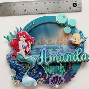 May include: A teal blue and purple cake topper featuring a mermaid with red hair and a green tail, a seashell, and the name "Amanda" in iridescent letters. The background is a blue and green ocean scene with a castle in the distance.