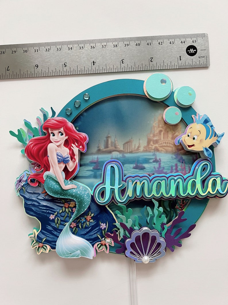 May include: A teal blue and purple cake topper featuring a mermaid with red hair and a green tail, a seashell, and the name "Amanda" in iridescent letters. The background is a blue and green ocean scene with a castle in the distance.
