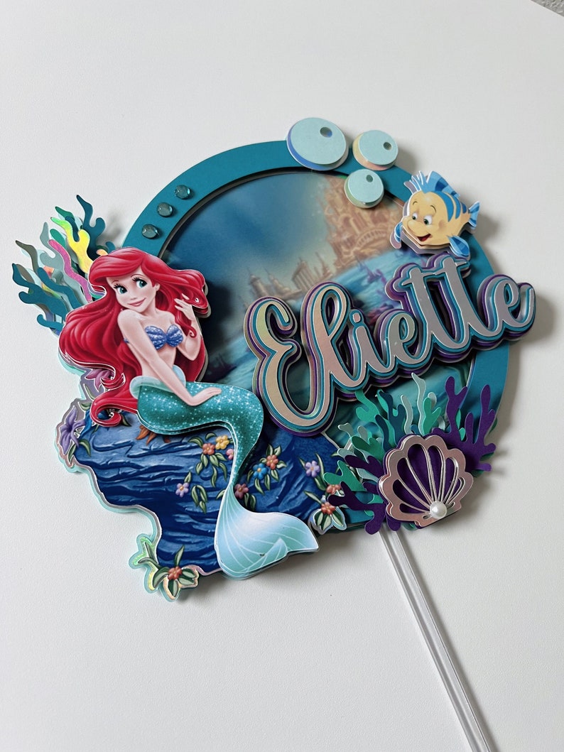 May include: A cake topper featuring Ariel, the Little Mermaid, sitting on a rock with her tail in the water. The topper also includes a blue and green seashell, coral, and the name "Eliette" in a colorful, layered font.
