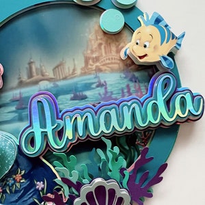 May include: A teal blue circle with a scene of a castle and boats in the ocean. The name Amanda is written in iridescent letters with a 3D effect. There are green and purple seaweed and a purple seashell with a pearl in the bottom right corner. A yellow fish with a blue stripe is in the top right corner.