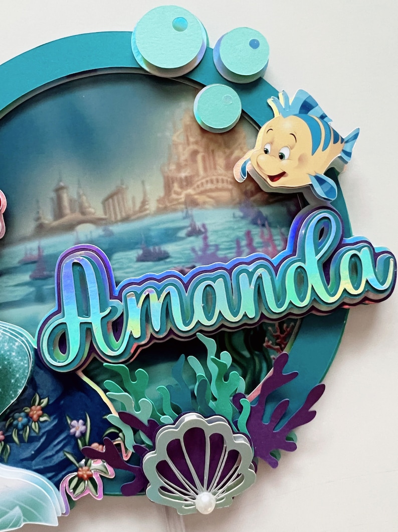 May include: A teal blue circle with a scene of a castle and boats in the ocean. The name Amanda is written in iridescent letters with a 3D effect. There are green and purple seaweed and a purple seashell with a pearl in the bottom right corner. A yellow fish with a blue stripe is in the top right corner.