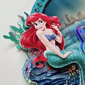 May include: Ariel, the Little Mermaid, sits on a rock with a blue and green tail and a blue seashell bra. She has long red hair and is smiling. The background is a blue ocean with a city in the distance.
