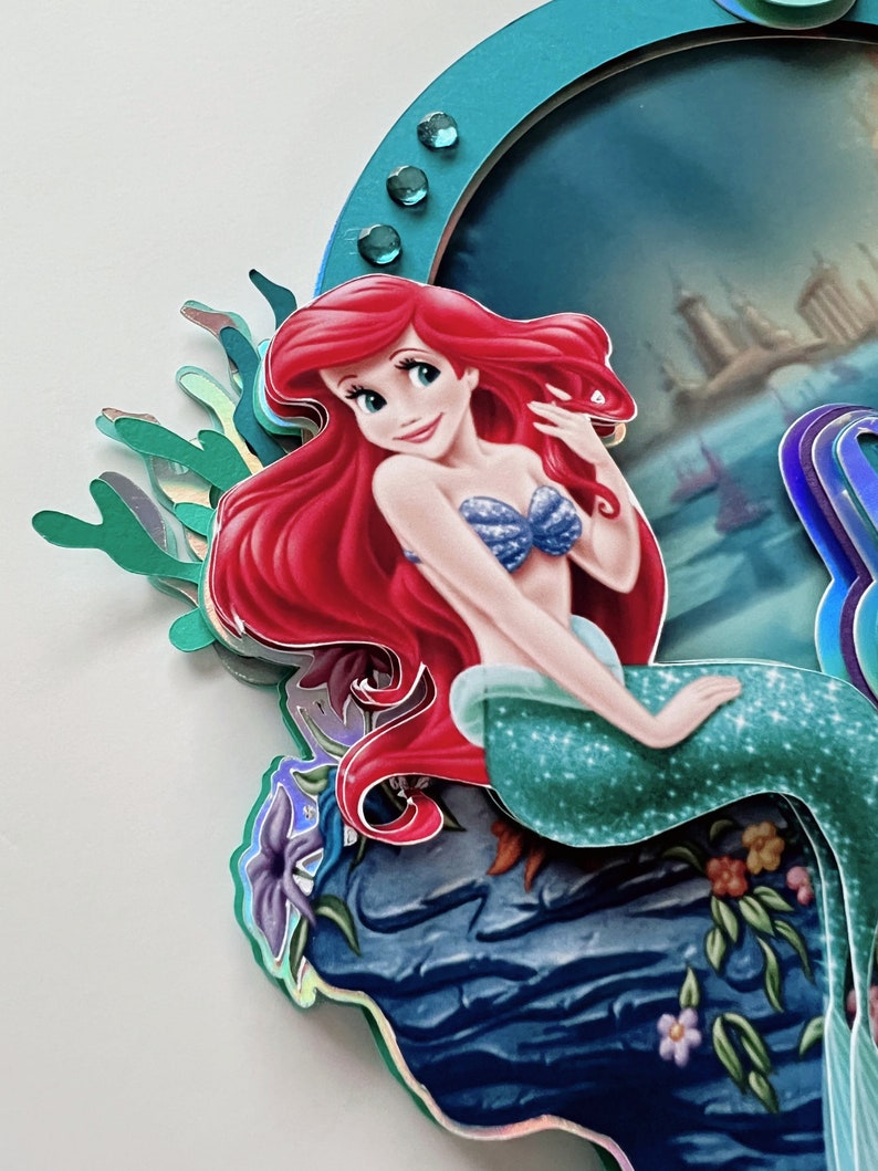 May include: Ariel, the Little Mermaid, sits on a rock with a blue and green tail and a blue seashell bra. She has long red hair and is smiling. The background is a blue ocean with a city in the distance.