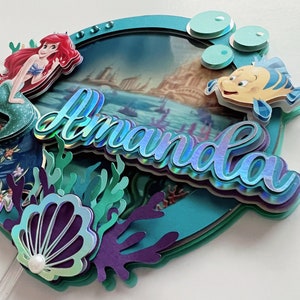 May include: A teal blue cake topper with a mermaid, a seashell, coral, and the name "Amanda" in iridescent letters. The background features a scene from the Little Mermaid movie.