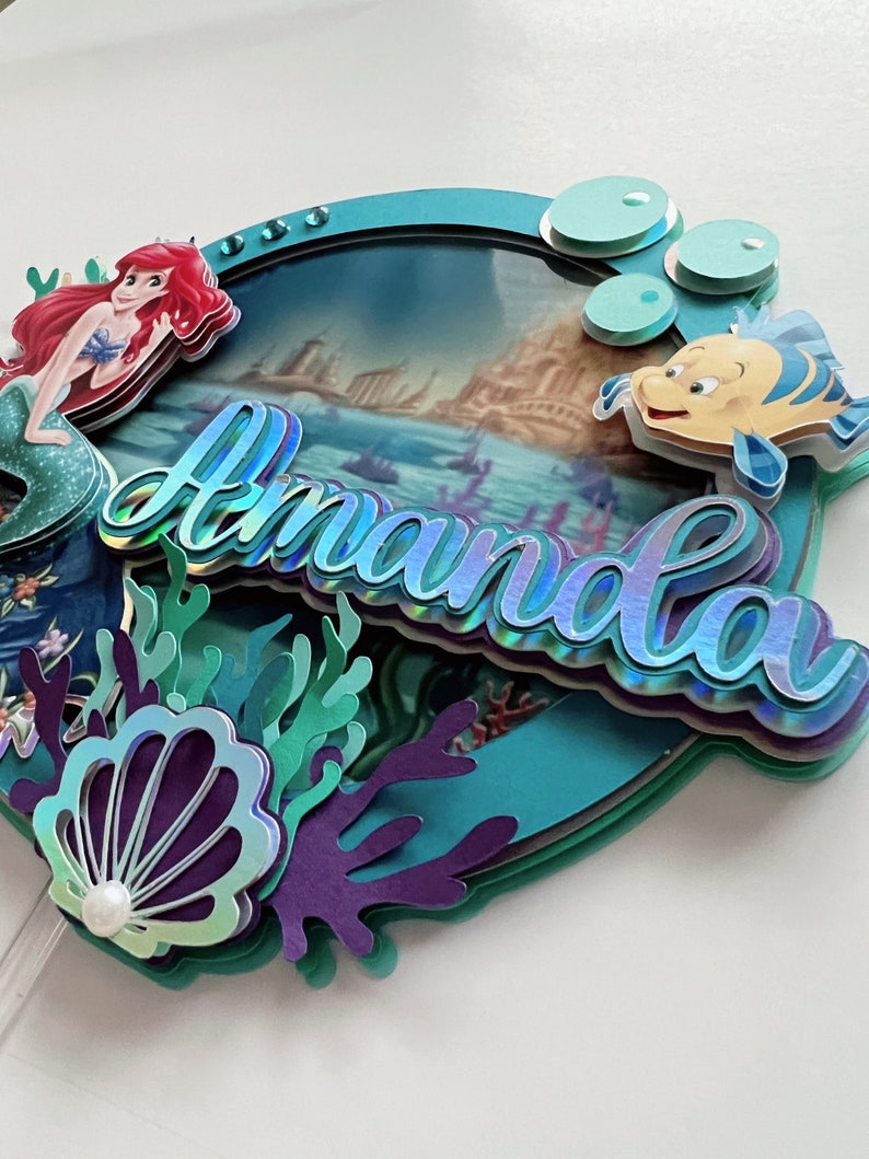May include: A teal blue cake topper with a mermaid, a seashell, coral, and the name "Amanda" in iridescent letters. The background features a scene from the Little Mermaid movie.