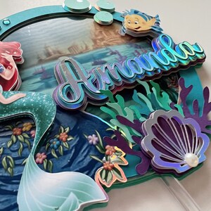 May include: A mermaid cake topper with a blue and green tail, a red and white top, and a picture of Ariel in the background. The topper also features a blue and green seashell, coral, and the name "Amanda" in iridescent letters.