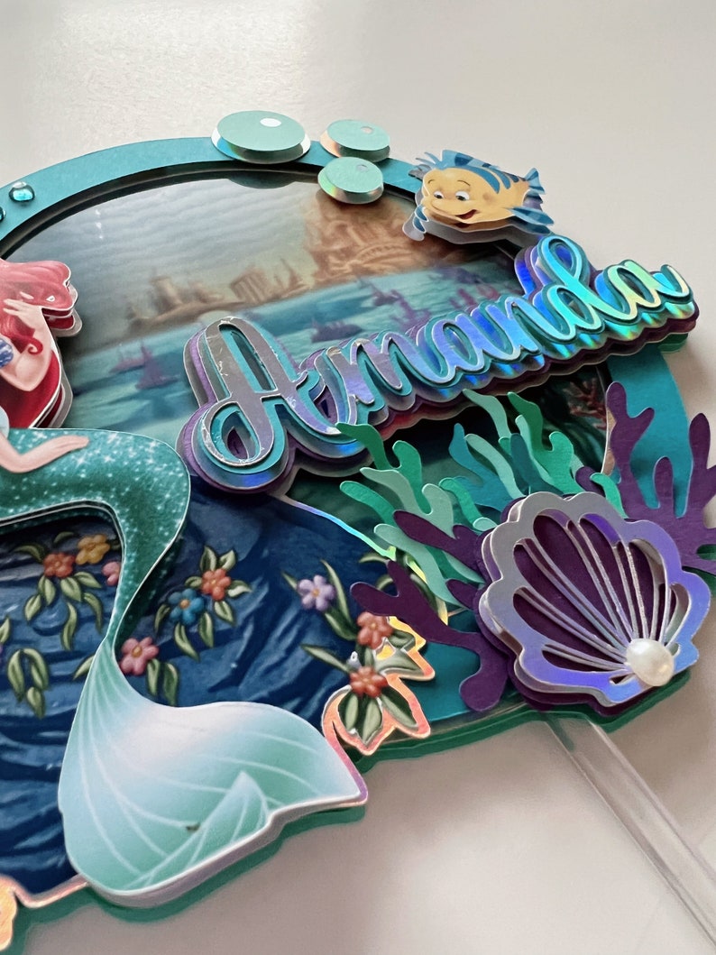 May include: A mermaid cake topper with a blue and green tail, a red and white top, and a picture of Ariel in the background. The topper also features a blue and green seashell, coral, and the name "Amanda" in iridescent letters.