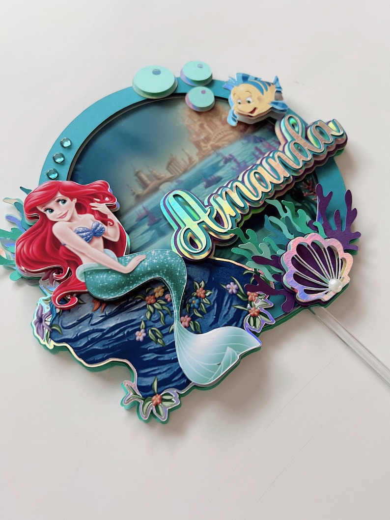 May include: A cake topper featuring Ariel, the Little Mermaid, with a blue and green mermaid tail, red hair, and a purple seashell. The topper also includes a blue and green ocean scene with a fish and the name "Amanda" in iridescent letters.