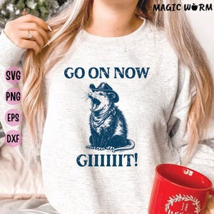 Może przedstawiać: Jasnoszara bluza z granatową grafiką oposa w kapeluszu kowbojskim i chuście, z napisem "GO ON NOW GIIIIIT!". Czerwony kubek widoczny w prawym dolnym rogu.