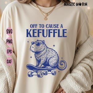 May include: Cream-colored t-shirt with a blue graphic of a capybara wearing sunglasses on a skateboard. The text "OFF TO CAUSE A KEFUFFLE" is above the capybara. The words "SVG, PNG, EPS, DXF" are on the left side.