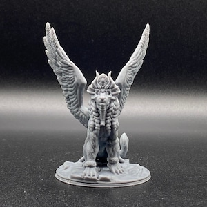 May include: A grey 3D printed miniature of a winged lion with a crown and a feathered headdress. The lion is standing on a round base.