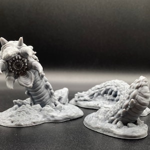 May include: A gray 3D printed miniature of a worm-like creature with a large mouth and sharp teeth. The creature is posed as if it is emerging from the ground.