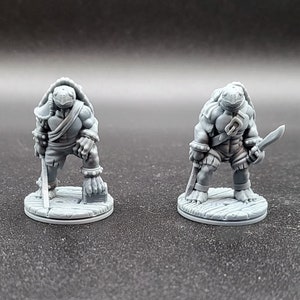 May include: Two gray painted miniature figurines of turtle-like creatures with swords. The figures are standing on round bases.