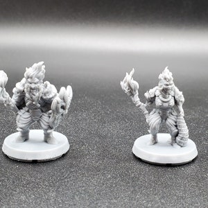May include: Two grey painted miniature figurines of fantasy characters. The figures are standing on round bases. The figures are both wearing armour and holding weapons. One figure is holding a staff with a flame at the top. The other figure is holding a hammer with a flame at the top.